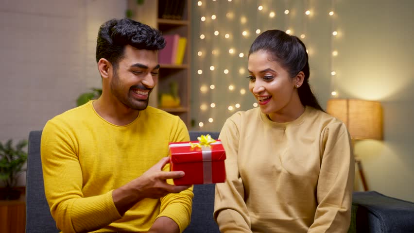 Happy young man giving surprise gift to girlfriend during wedding anniversary on sofa at home - concept of affection, dating and relationship Royalty-Free Stock Footage #1099992743