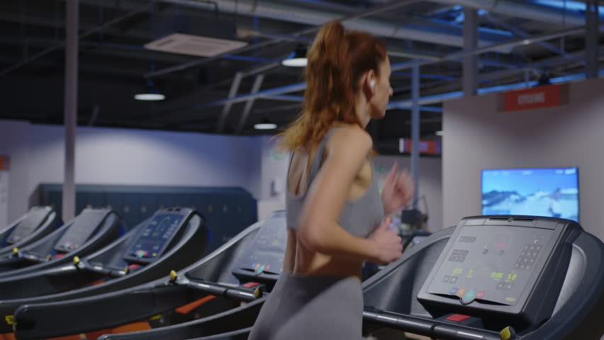 Rear view, a girl with long hair in headphones runs on a treadmill in a modern gym. The concept of a healthy lifestyle and taking care of your body