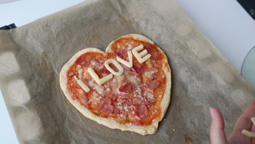 Woman hand hand lays out the inscription from cheese I love you. Italian heart shaped pizza with salami, tomato sauce, parmesan, sauce, mozzarella and olive oil on parchment paper. Love concept Royalty-Free Stock Footage #1099997677