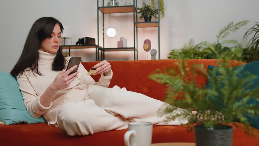Young brunette woman using credit bank card and smartphone while transferring money, purchases online shopping, order food delivery at home apartment indoors. Happy girl in living room lying on couch | Shutterstock HD Video #1099999611