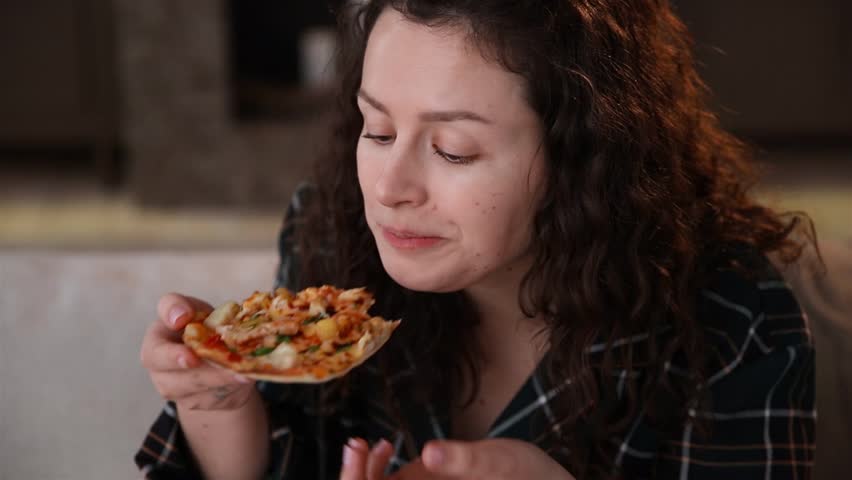 Young dark-haired curly caucasian girl enjoys pizza at home on the sofa on her weekend. Woman celebrating the end of the diet by ordering a delicious pizza home and eating it in her pajamas | Shutterstock HD Video #1100000561