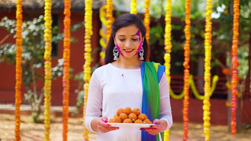 Happy smiling girl with coloured face holding sweet or laddu by looking at camera on flower decorated background - concept of holi festival, happiness and joyful. Royalty-Free Stock Footage #1100001727