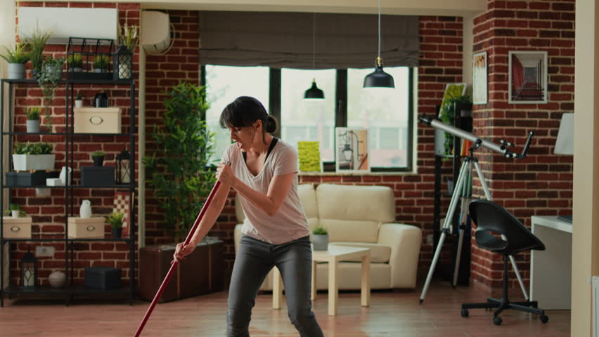 Smiling person cleaning living room floors with mop, listening to music and doing house chores. Cheerful woman dancing and washing dirt, doing housework in apartment. Spring cleaning. Royalty-Free Stock Footage #1100004925