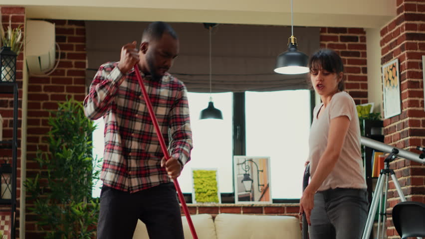 Diverse life partners dancing and singing with appliances, using mop and vacuum cleaner to wash apartment floors. Young couple having fun with music while they clean household. Handheld shot.