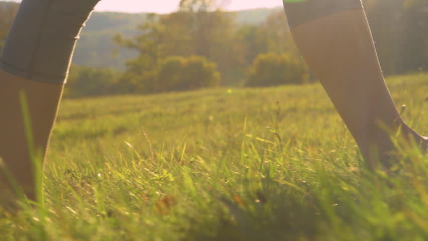 CLOSE UP, LOW ANGLE: Barefoot young woman walking on green grass in golden light. Carefree and relaxing moment in nature on a beautiful sunny morning. Female person pacing the meadow with bare feet. Royalty-Free Stock Footage #1100010719