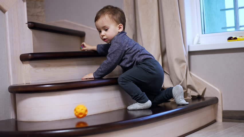 Adorable baby sitting on the stairs indoors. Little toddler watching the ball roll down and follows it.