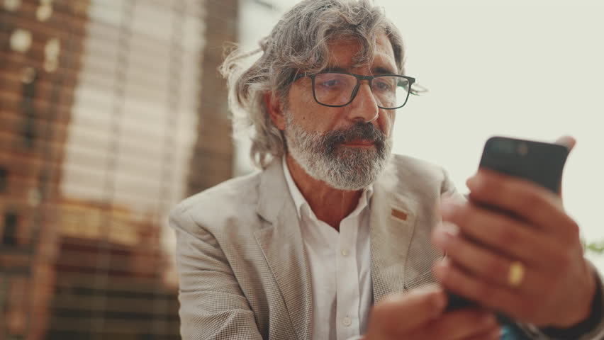 Clouse up, mature businessman with beard in eyeglasses wearing gray jacket is using cell phone. Middle aged manager scrolling information on his smartphone while sitting outside the office | Shutterstock HD Video #1100016739