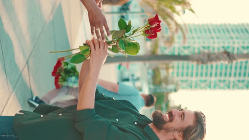 VERTICAL VIDEO, Close-up of young happy man and woman joyfully hugging when meeting on the embankment, man gives girl a bouquet of red roses. Closeup, joyful date of young couple | Shutterstock HD Video #1100016787