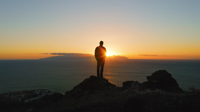 Man standing on top of rock with epic mountain viewpoint colorful sunset light and endless ocean horizon. Drone aerial landscape.