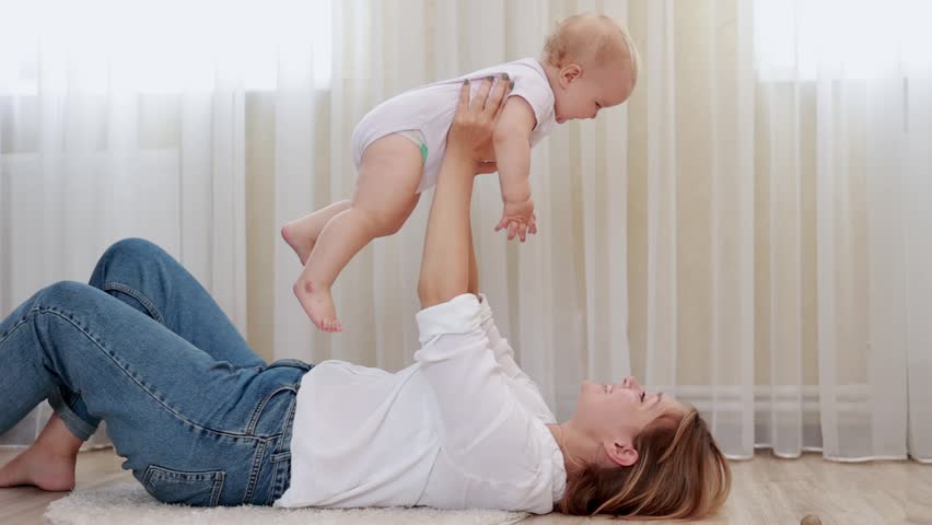 Happy loving millennial caucasian mom lie on back on floor in light cozy bedroom lift up toss in air small baby daughter or son.Caring mum laugh cuddle kiss cute infant babe have fun enjoy motherhood. | Shutterstock HD Video #1100018869
