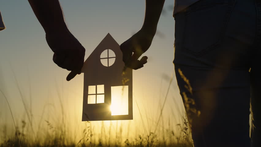 Familys hands are holding paper house at sunset, sun is shining through window. Symbol of house, happiness. Concept of building house for family. Dream to buy house. Real estate insurance. Family Home Royalty-Free Stock Footage #1100019673