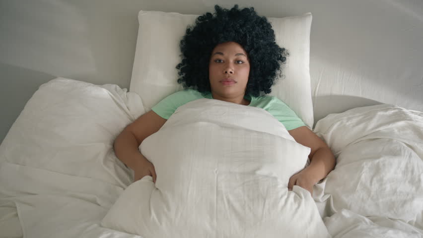 Top down view on young African American woman lies on bed and covering her head by large pillow hiding from reality, do not want to see the light and wake up. Depression or mental health problems shot Royalty-Free Stock Footage #1100023695