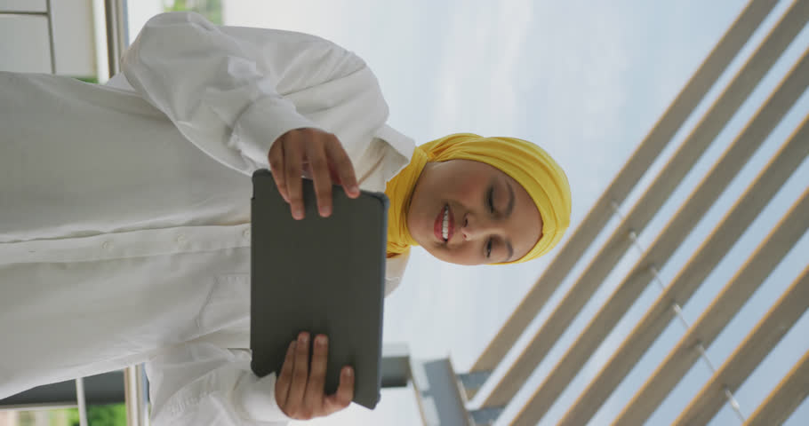 Vertical video of happy biracial businesswoman celebrationg and using tablet at office. Working at modern office, business and finances concept.