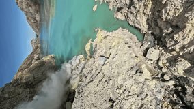 Dive to volcano crater island mountain rock crack stone sharp with acid lake turquoise blue water miss smoke fog aerial view. FPV sport drone shot pond cliff rocky terrain nature. Vertical format 4k