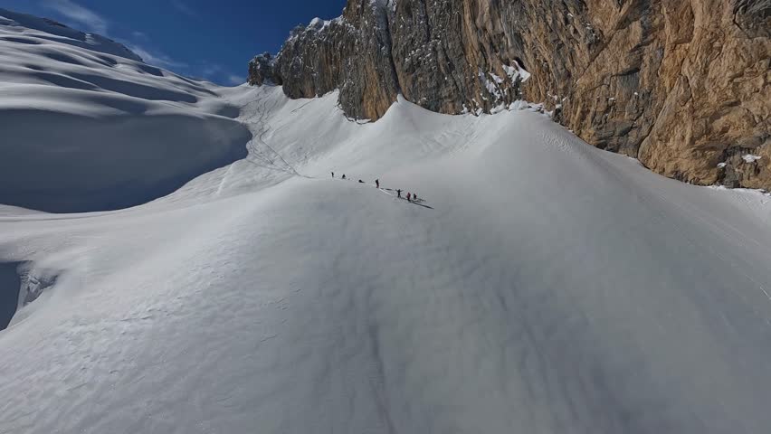 Ski tour heliski group standing ob big rock wall. Extreme sport tourist skiing snowboarding break riding snow mountain aerial view FPV drone shot travel expedition freeride winter cliff valley scenery