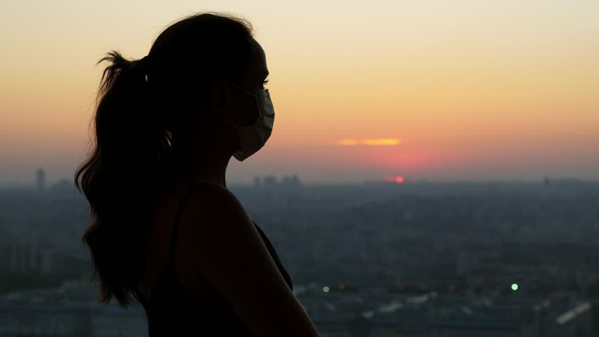 Woman in quarantine, standing in a medical mask in semi-darkness against the background of sunset and city from above. Hand-held shot from back, blurred background | Shutterstock HD Video #1100026965