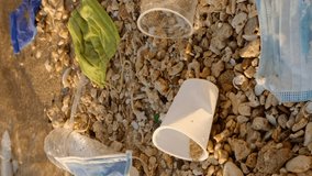 VERTICAL VIDEO, Close-up of disposable plastic, face mask and other debris on corals beach in surf zone. Plastic and other garbage on the seashore. Slow motion, Camera moves sideway to the left side