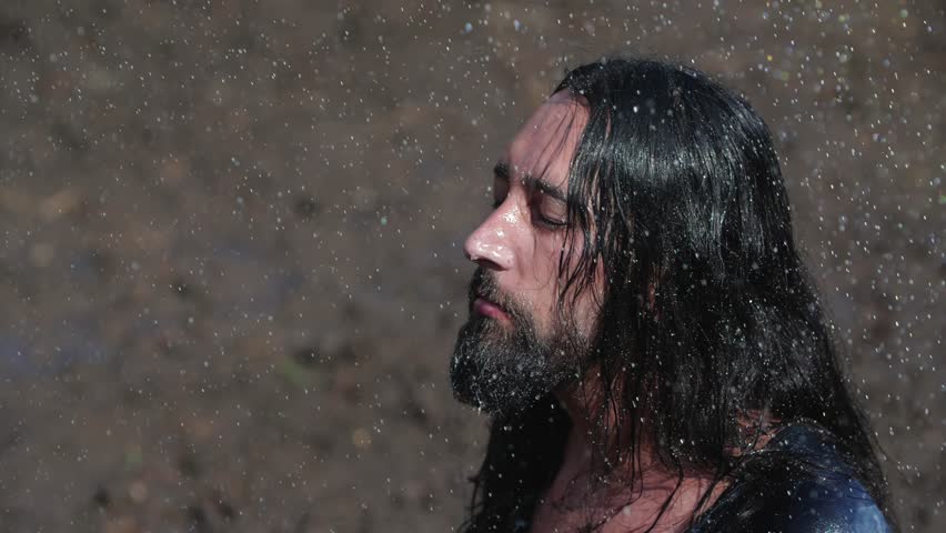 Close up wanderer rejoices in the long-awaited rain in the desert. An emaciated man with long dark hair stretches out his arms to the sky as raindrops fall on him. 200 fps slow motion