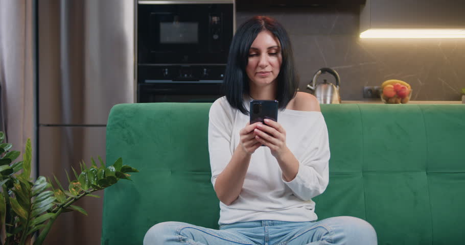 Happy young woman sitting on a green sofa and using a smartphone smiling in a modern kitchen. Female holding phone texting message technology app, typing email at home. | Shutterstock HD Video #1100034003