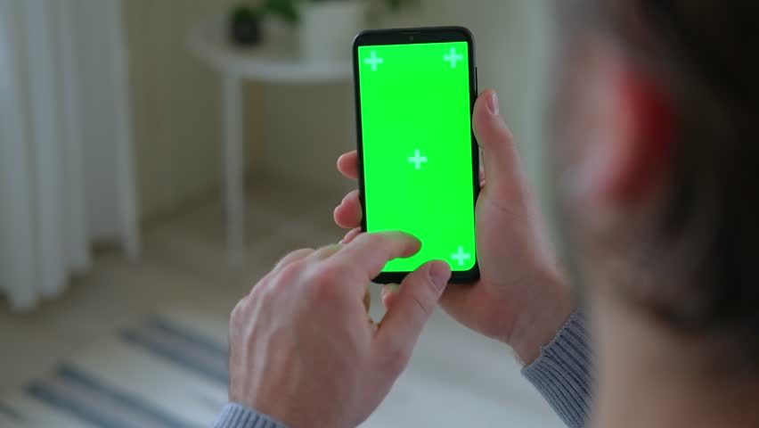 Young man sitting at home holding smartphone green mock-up screen in hand. Male person using chroma key mobile phone. Vertical mode. Touching, swiping display, tapping, surfing Internet social media | Shutterstock HD Video #1100035293