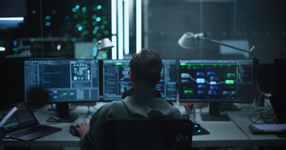 Lone Hacker Breaks Into Government Data Servers and Infects Their System with an Exploit Software. His Hideout Place is in Dark Technological Facility with Multiple Displays. Footage from the Back Royalty-Free Stock Footage #1100036235