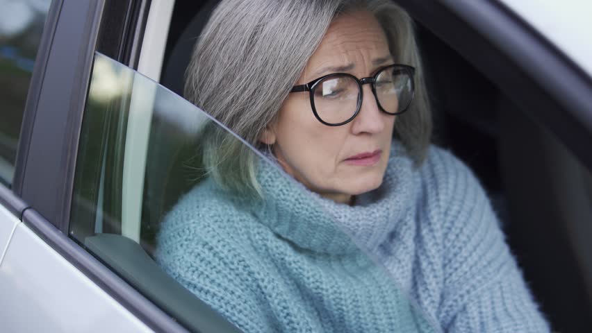 Unhappy mature woman in elegant eyeglasses and cozy warm sweater sitting on passenger seat of automobile, arrested by police for misbehavior or expired driving license