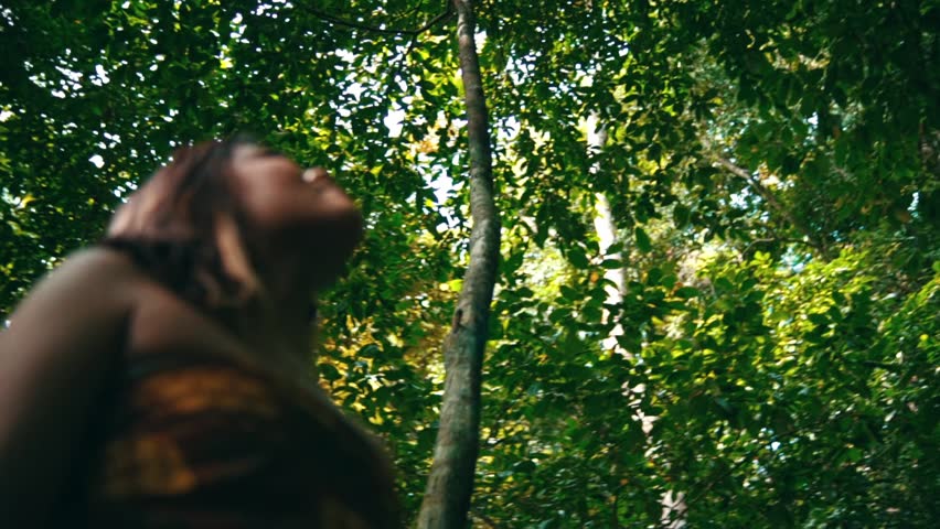 Low-angle panning shot with a woman out-of-focus in the foreground, looking up at the tall trees and bright sun in a lush green forest. Ethereal and dreamy