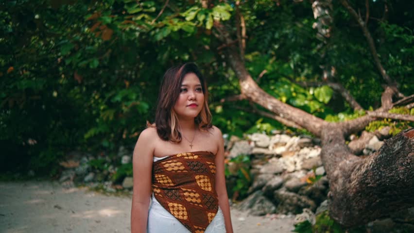 A beautiful Asian woman in traditional attire walks barefoot on sand towards a large, fallen tree. She reaches up to touch the trunk, smiling in the sunlight.