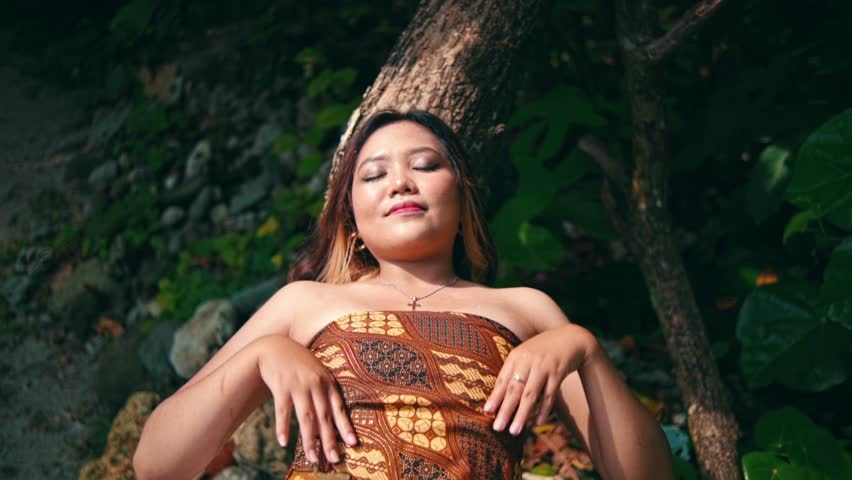 A serene Asian woman in a batik top leans against a tree with eyes closed. She gracefully raises her arms in a peaceful, meditative motion in a dark forest.