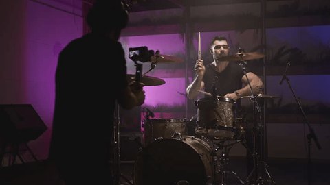 Shooting a music video. The director of photography makes a video, shoots a drummer musician. The view behind the scenes. Create a music video for a rock band. Creative director and crew. Adlı Stok Video