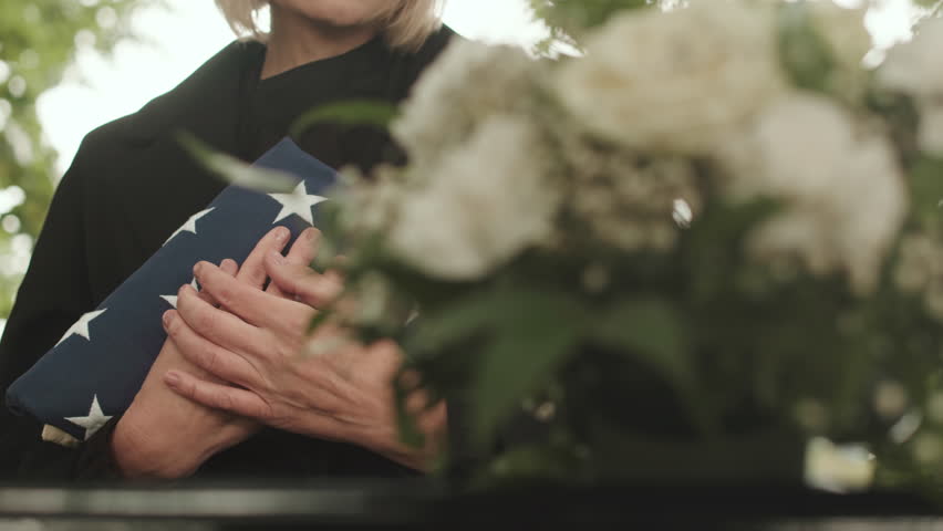 Cropped shot of unrecognizable blonde woman in black clothes holding US burial flag close to her chest while standing by closed casket at outdoor funeral ceremony