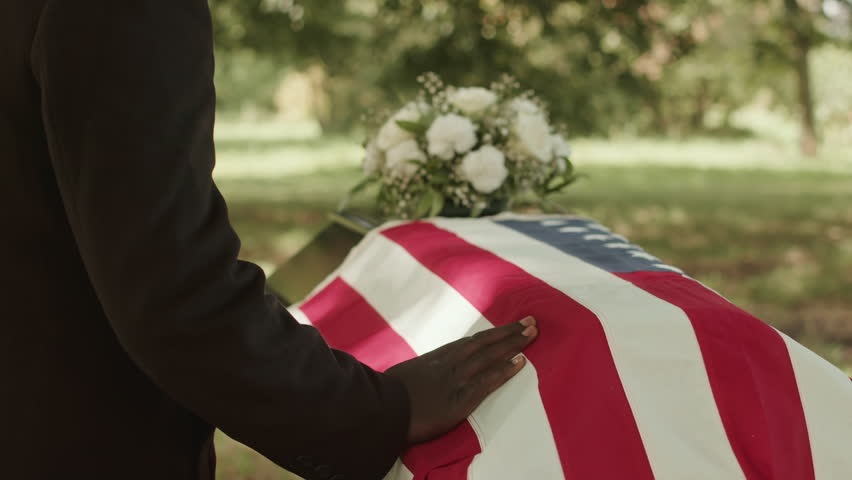 Unrecognizable Black male hand sliding on casket lid with unfolded US burial flag on it outdoors at funerals