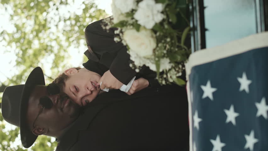 Vertical shot of multiracial couple standing by casket at funeral ceremony mourning