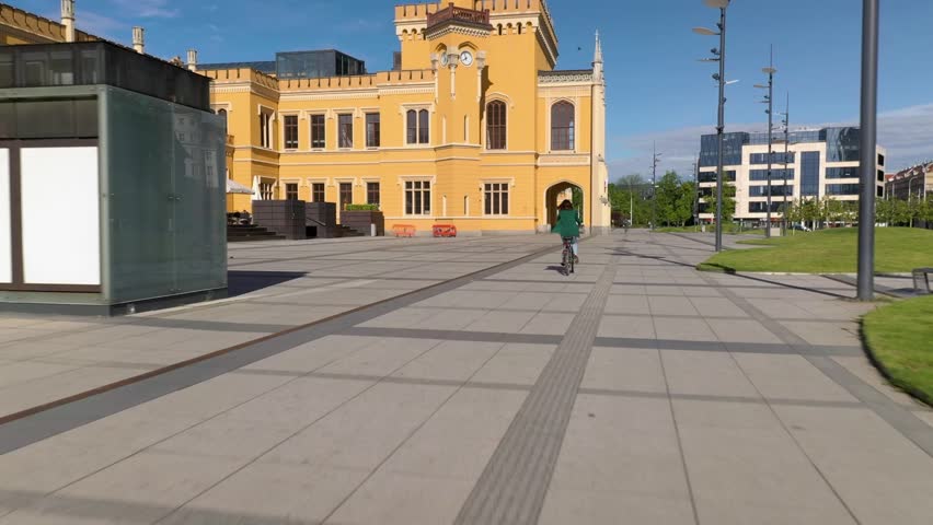 Young girl with green jacket, riding a bike next to the orange renovated building in the city. Drone follows her at low altitude. 