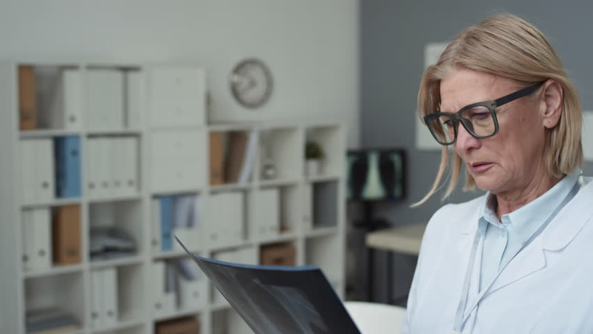 Serious mature female radiologist in eyeglasses and lab coat looking attentively at x-ray image of lungs of patient by her workplace while working in medical office | Shutterstock HD Video #1100046487