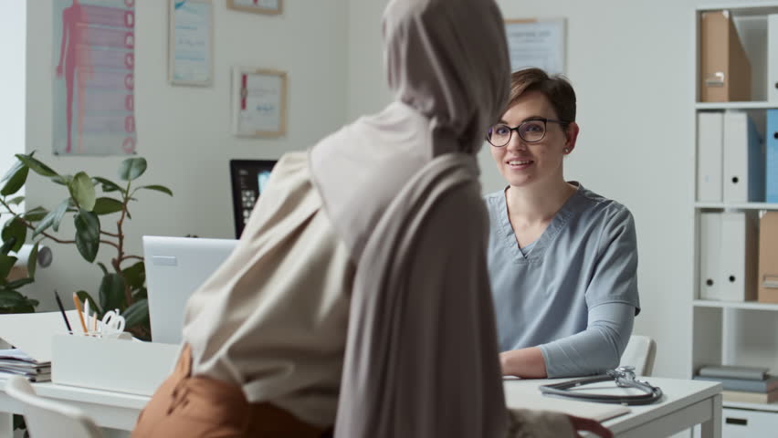 Young confident clinician in uniform consulting sick Muslim female patient in hijab sitting down by desk in front of her, describing symptoms and complaining about feeling unwell Royalty-Free Stock Footage #1100047567