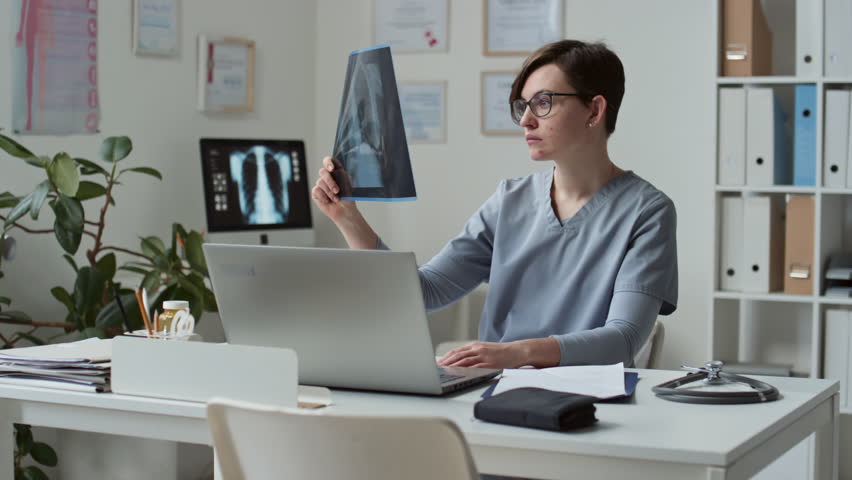 Young serious female clinician in eyeglasses and blue medical scrubs learning x-ray image of patient during online consultation while sitting by workplace in office | Shutterstock HD Video #1100050219