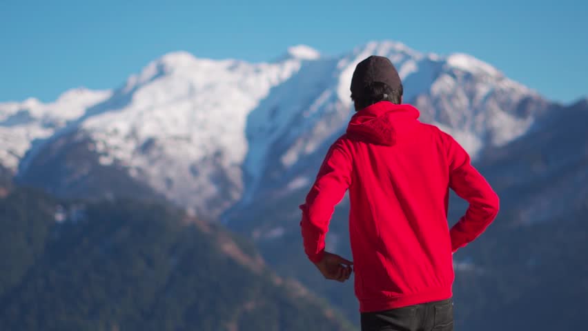 Rear view of an Indian man wearing red hoodie staring at the snow covered mountains of Manali after snowfall in winter season at Himachal Pradesh, India. Tourist looking at the snow covered mountains.
