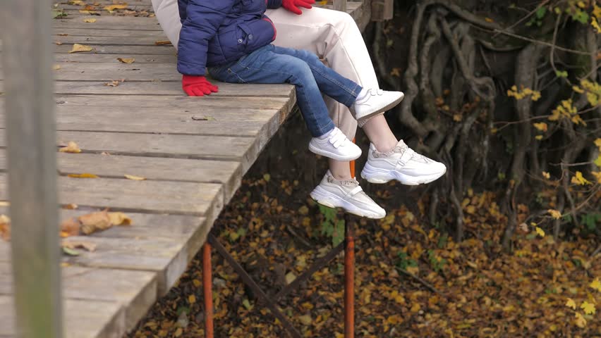 Close up video of young mother and son are sitting on river bridge and looking at water flow. They spend leisure time together. A woman and 2 years old boy sit on a wooden bridge.Day vacation on river