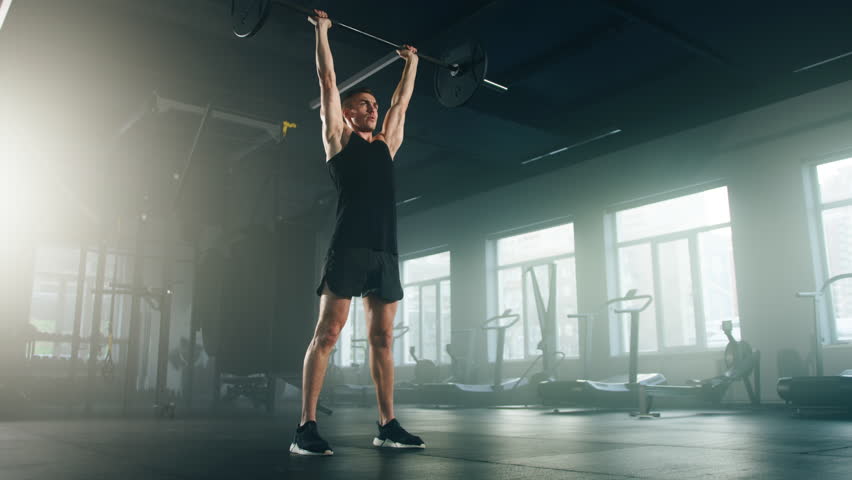 Close-up view of sporty, young bodybuilder lifting a barbell with loaded heavy weights. Active sportsman in black sportswear having intensive training indoors. High quality 4k footage