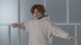 Curly young man enjoying music and dancing in studio. Smiling dancer showing his moves and pointing with finger to the camera. Guy in hoodie recording dance tutorial
