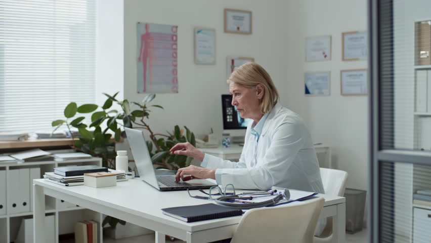 Mature radiologist sitting by workplace in front of laptop, pressing button on keyboard, taking lung x-ray of online patient and looking at image during remote consultation | Shutterstock HD Video #1100061037