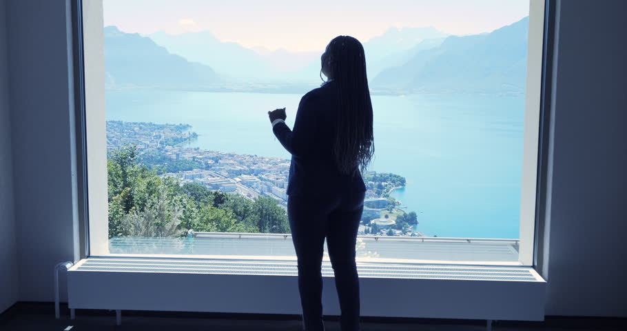 Succesful Female Sales Person on Business Travel Chatting on Mobile Phone While Looking Through Window at Lake city landscape