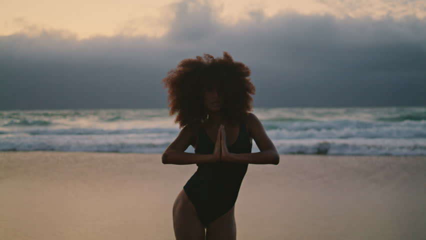 Silhouette of slim flexible woman performing belly dance on sand beach at overcast evening. African american attractive girl wearing black swimsuit making smooth body movements at evening seashore.