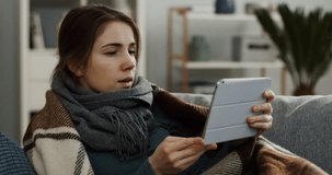 Close up of the unwell young woman in a scarf and a plaid having a videochat on the tablet device, caughing in a napkin as having a cold. Portrait shot. Indoors
