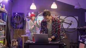 Beautiful caucasian young man using laptop online shopping for bike parts and tools with cute attractive woman fixing bike at modern cycling shop workshop. Lovely couple having business. - Powered by Shutterstock - Get 15% off with code: PIKWIZARD15