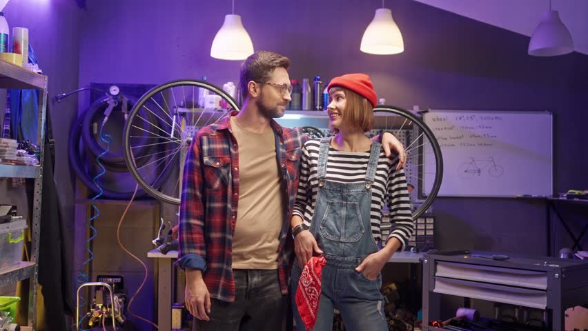 Portrait of two young caucasian beautiful people standing in modern workshop with neon lights looking at camera smiling hugging. Cute loving couple of mechanics spending time together.