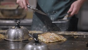 This slow motion video shows the hands of a chef flipping a okonomiyaki pancake on a grill.
