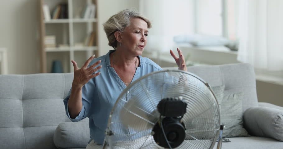 Overheated 60s woman sit in front of ventilator, cools herself, suffers from heat inside living room, feels badly, breath fresh air from electric fan, hot weather at summer day without air-conditioner Royalty-Free Stock Footage #1100081143