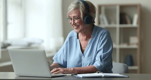 Happy senior woman in headphones sit at desk, enjoy on-line class with tutor, gain new knowledge, improve skills, noting, spend time on retirement use videocall app. Education of retiree at home, tech
